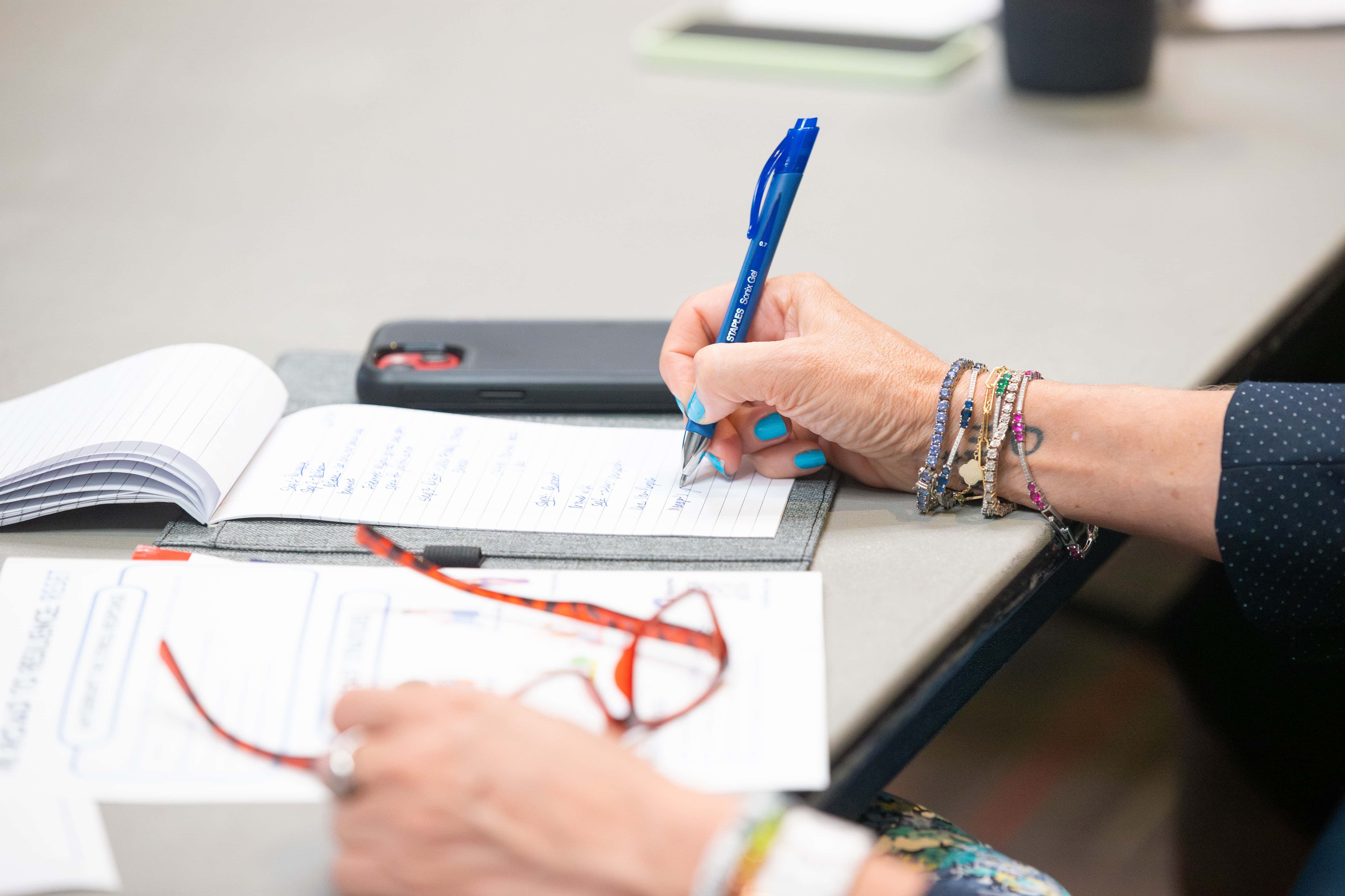 Woman taking notes with her pen in one hand writing on paper and her other hand holding her glasses.