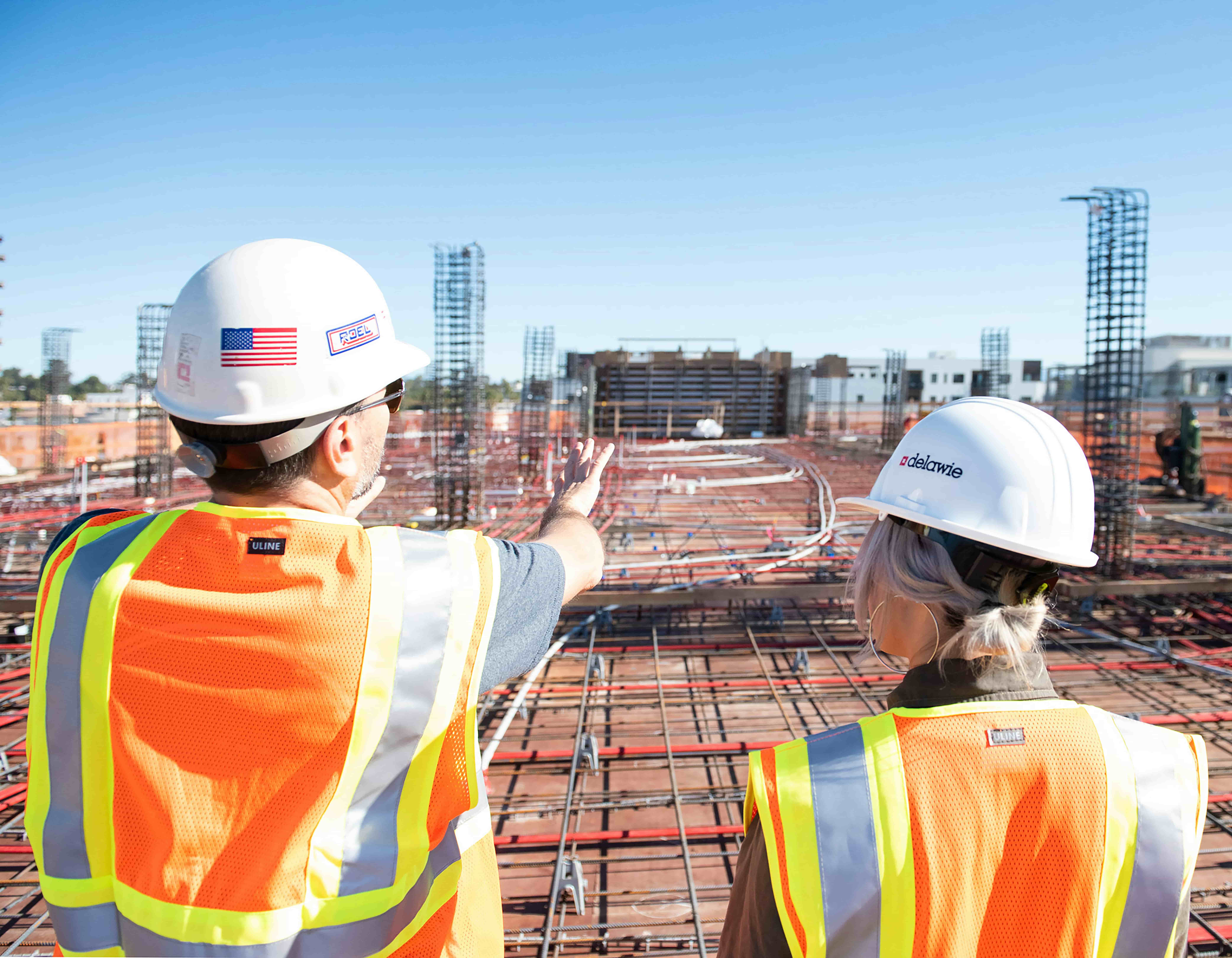 Two construction managers, male and female, wearing high-viz vests and helmets, overlooking a construction project.