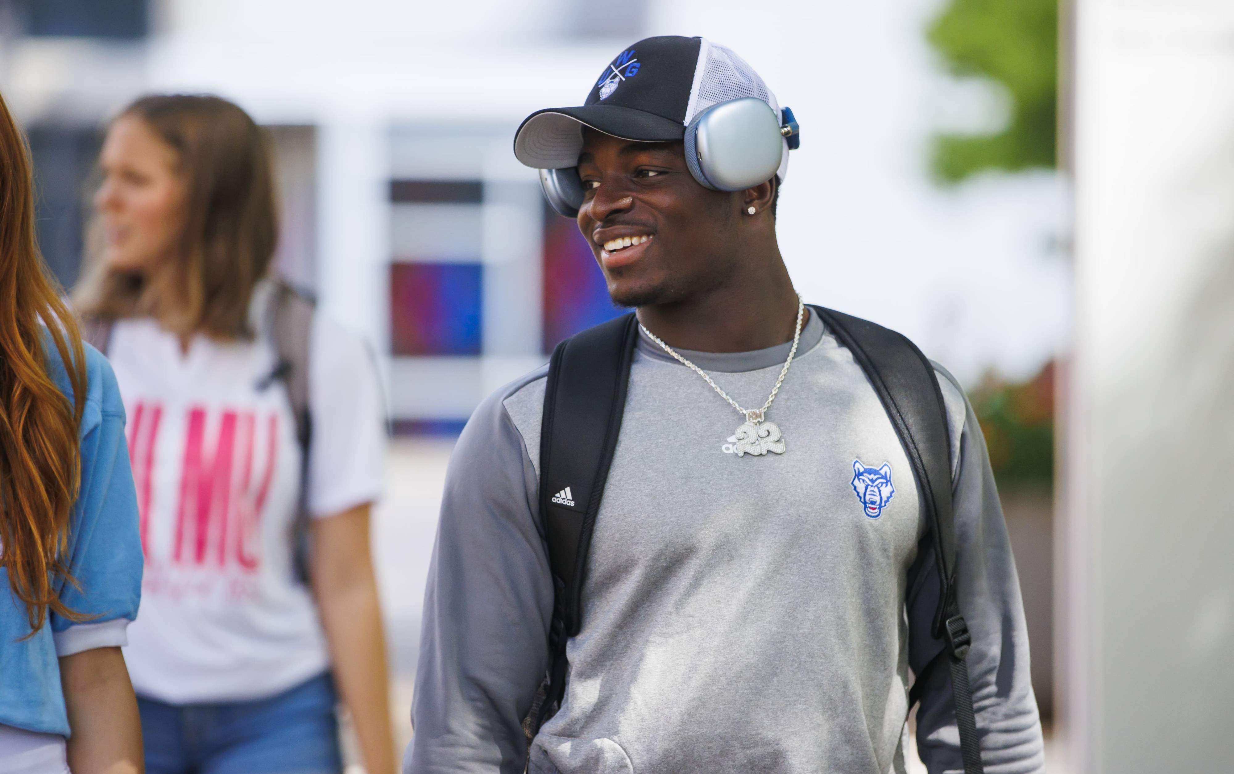 An African-American male student walking on campus smiling and wearing a UWG hat and long sleeve shirt.
